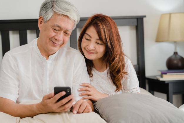 Asian senior couple using mobile phone at home. asian senior chinese  grandparents, husband and wife happy after wake up, watching movie lying on  bed in bedroom at home in the morning concept.